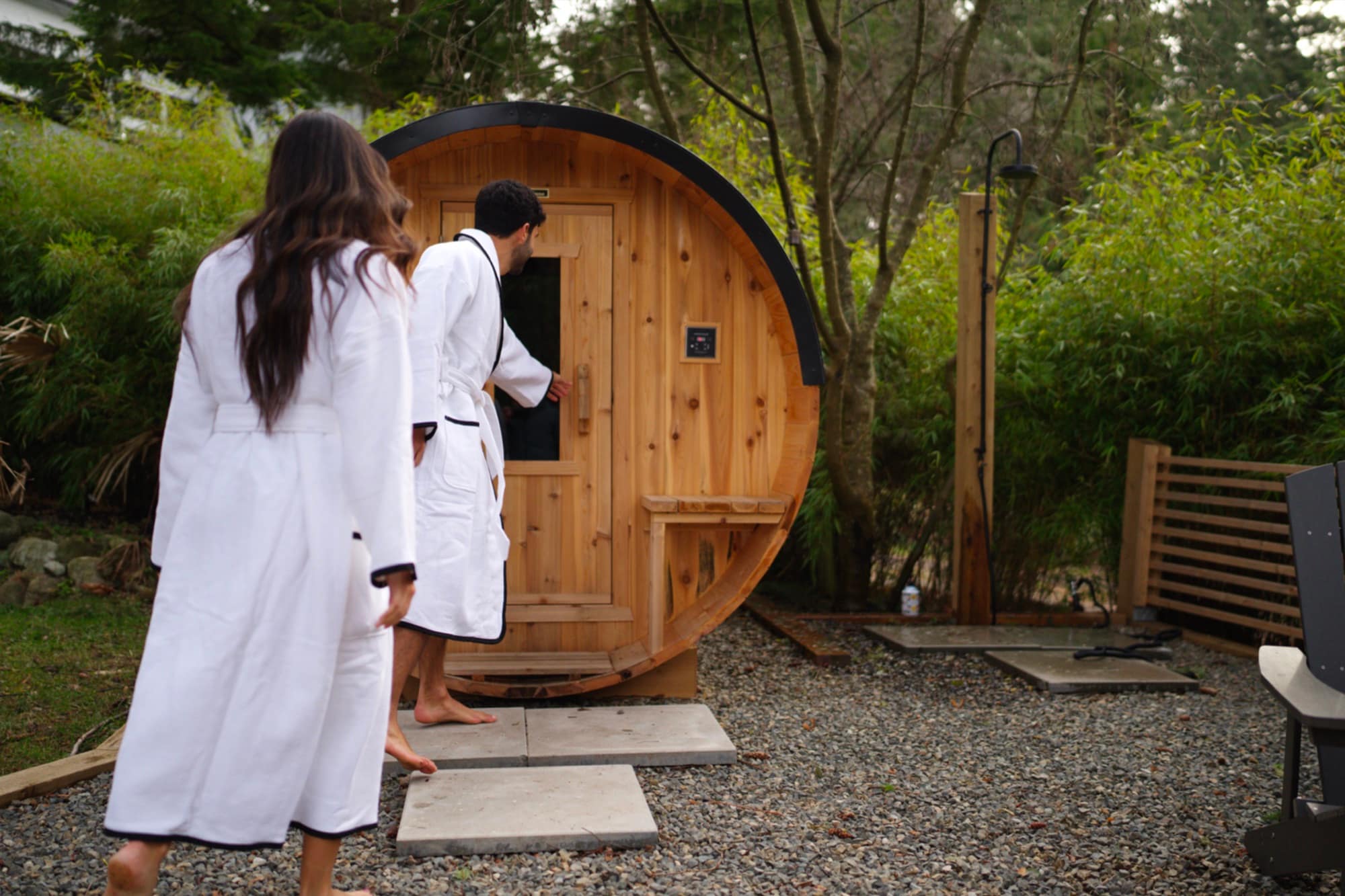 Two people in white robes stepping into a cedar Nootka barrel sauna beside an outdoor shower in a green backyard