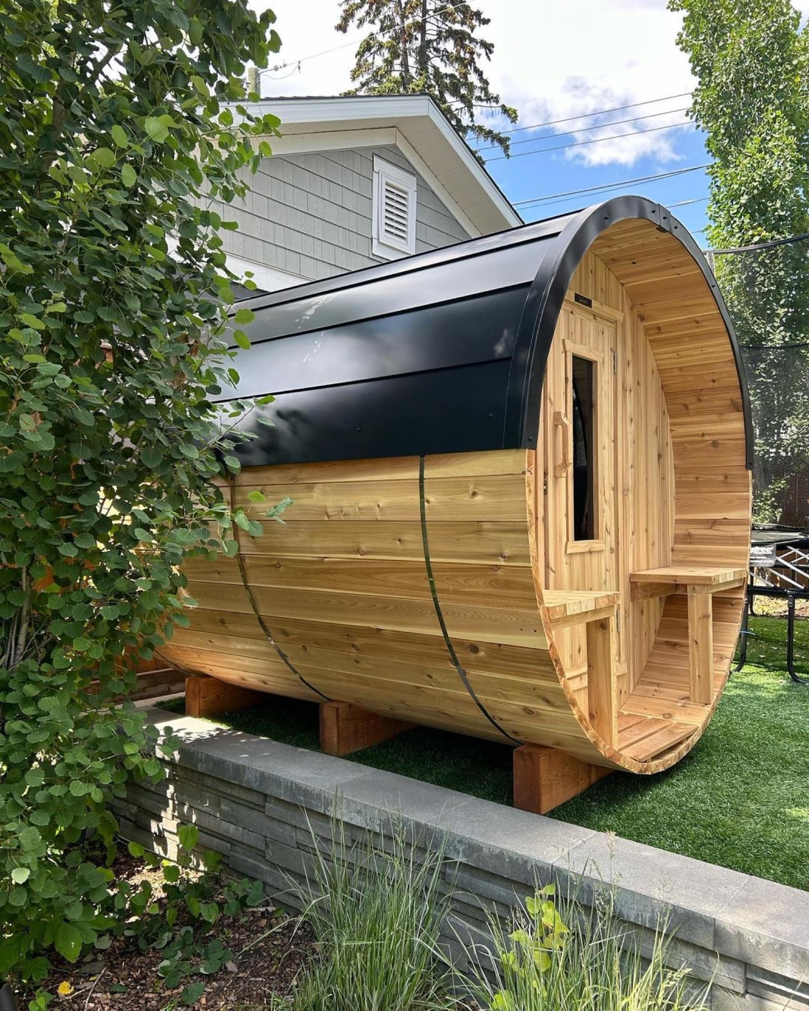 Cedar Nootka barrel sauna with a black steel roof on a raised garden bed in a residential backyard