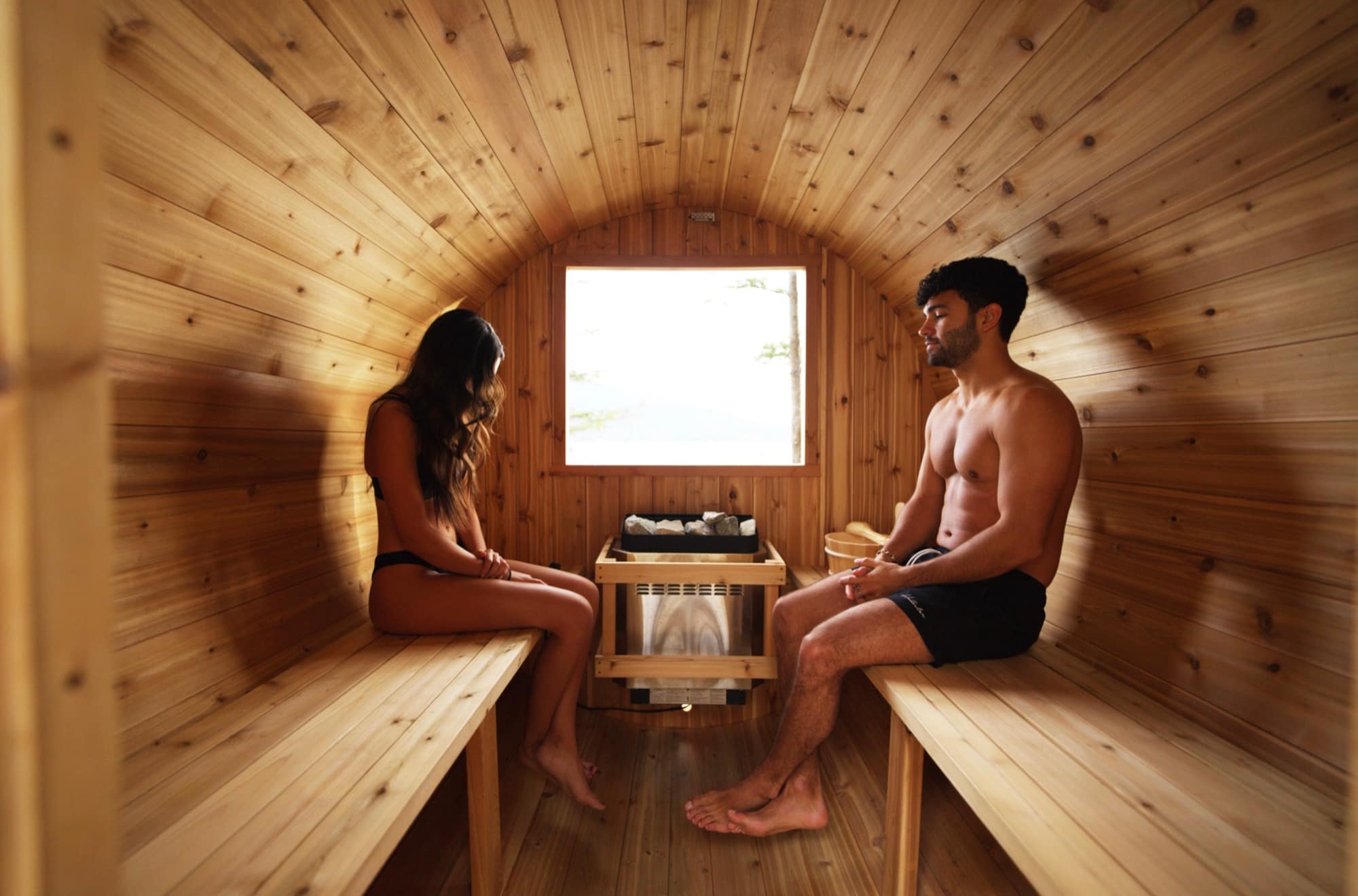 Interior of a Nootka cedar barrel sauna: two people on facing benches beside a stone-topped tower heater below a bright window