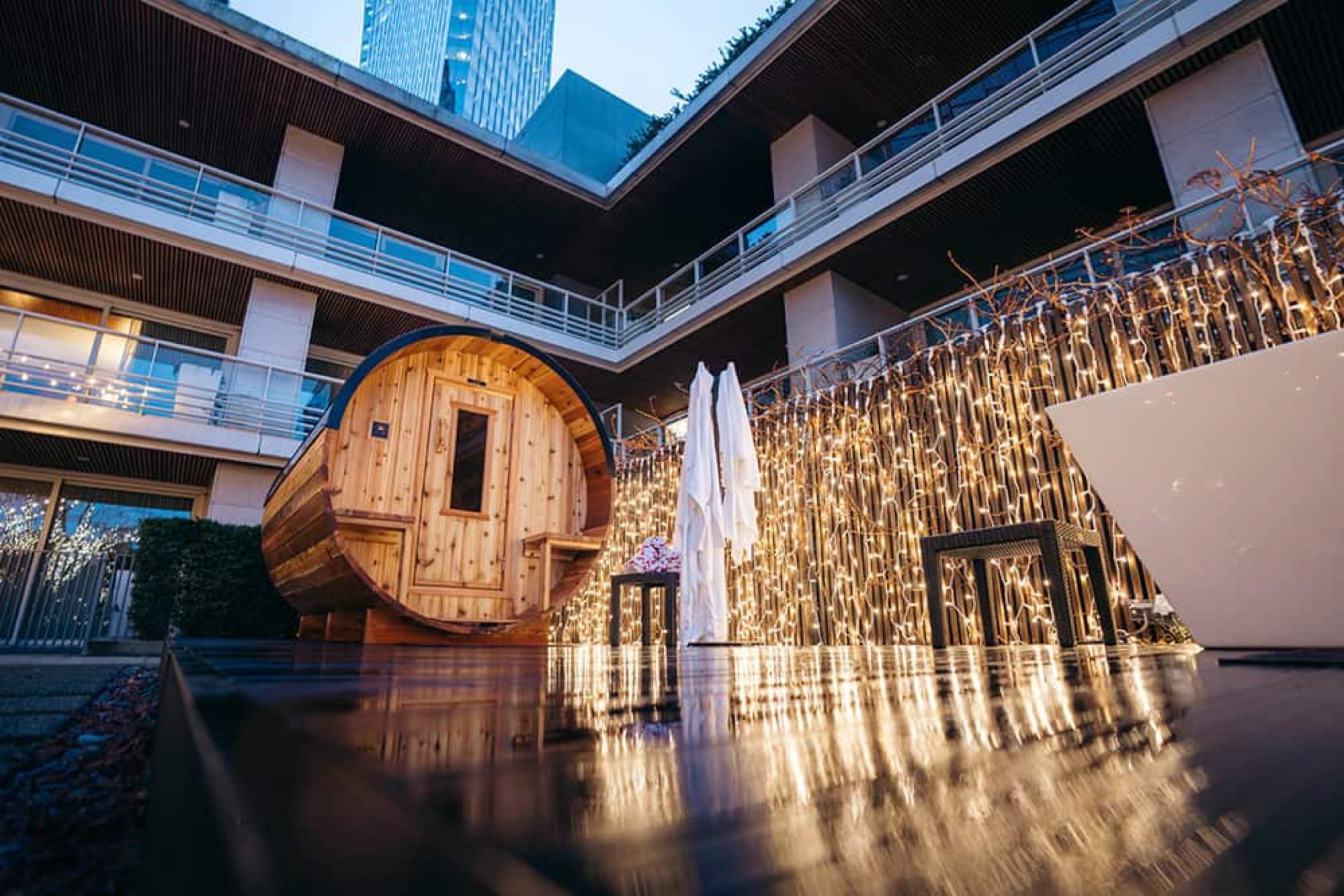 A Nootka barrel sauna in a luxury hotel courtyard strung with warm fairy lights beside a freestanding tub at dusk