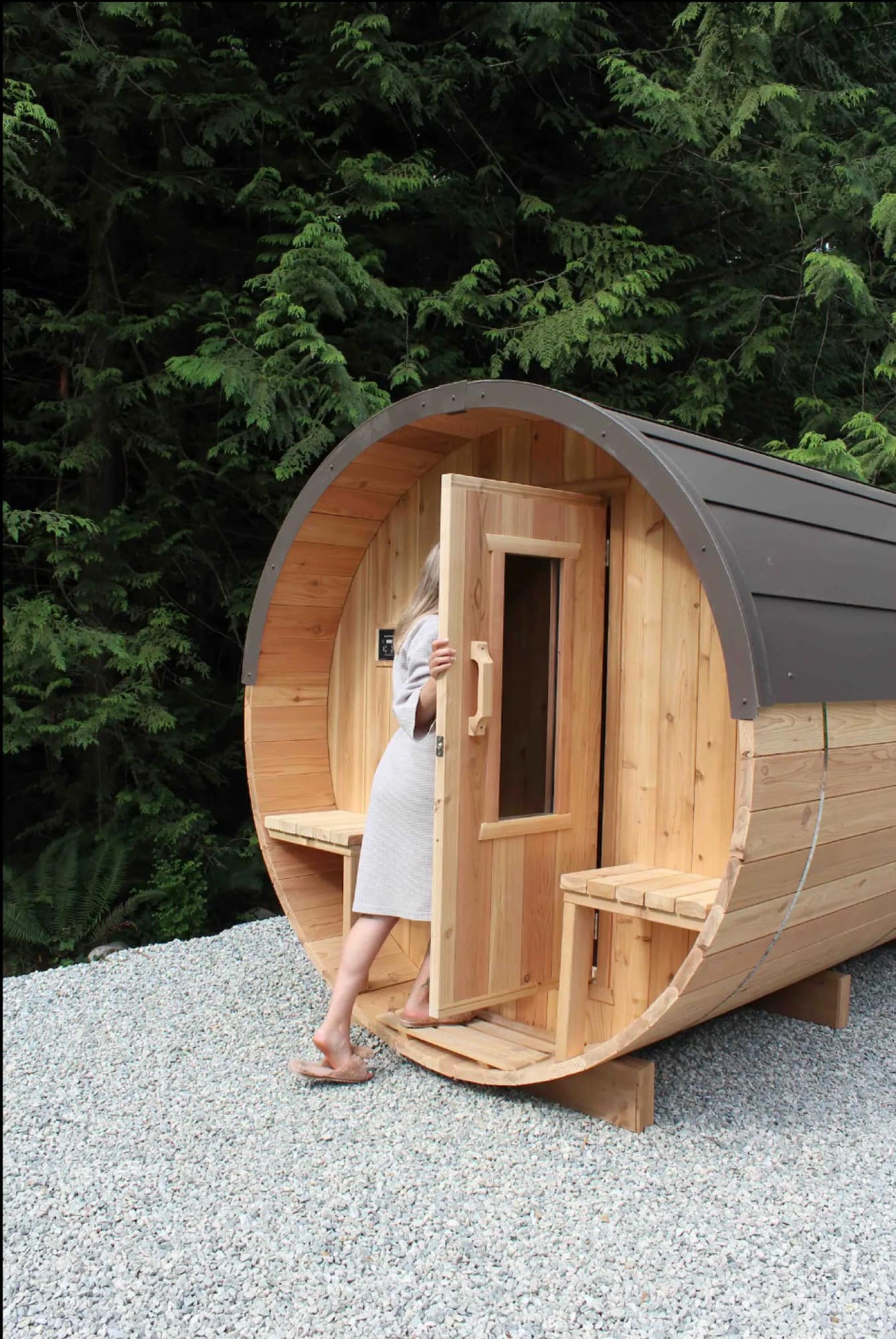A person stepping into a cedar Nootka barrel sauna set on gravel against a wall of deep-green West Coast forest